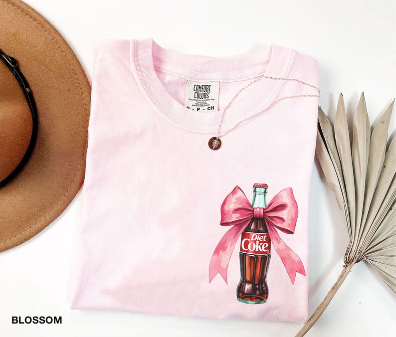 May include: Pink t-shirt with a graphic of a Diet Coke bottle with a pink bow. The shirt has a Comfort Colors tag.