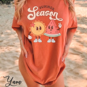 May include: A woman wearing a burnt orange t-shirt with a graphic of a smiling sun and watermelon with the text "Summer Season".
