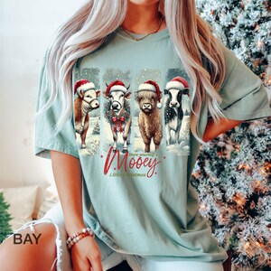 May include: A light green t-shirt featuring four cows wearing Santa hats, with the text "Have yourself a Mooey Little Christmas." The cows are depicted in a snowy winter scene, creating a festive holiday design.