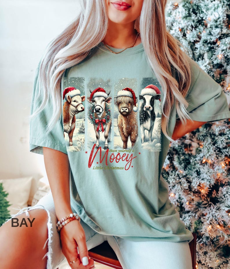 May include: A light green t-shirt featuring four cows wearing Santa hats, with the text "Have yourself a Mooey Little Christmas." The cows are depicted in a snowy winter scene, creating a festive holiday design.