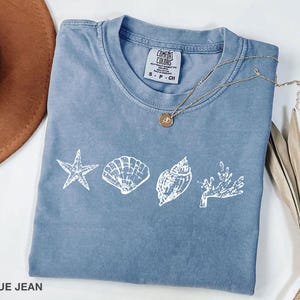 Op de afbeelding: Een blauw jeans T-shirt met een witte afbeelding van schelpen en een zeester. De tekst "COMFORT COLORS" is zichtbaar op het label.