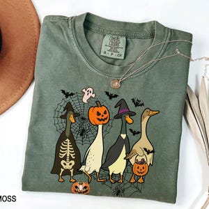Puede incluir: Camiseta Comfort Colors verde musgo con un dise&ntilde;o de Halloween. El dise&ntilde;o incluye cuatro patos disfrazados, una telara&ntilde;a, murci&eacute;lagos y un fantasma. Un collar plateado est&aacute; sobre la camiseta.
