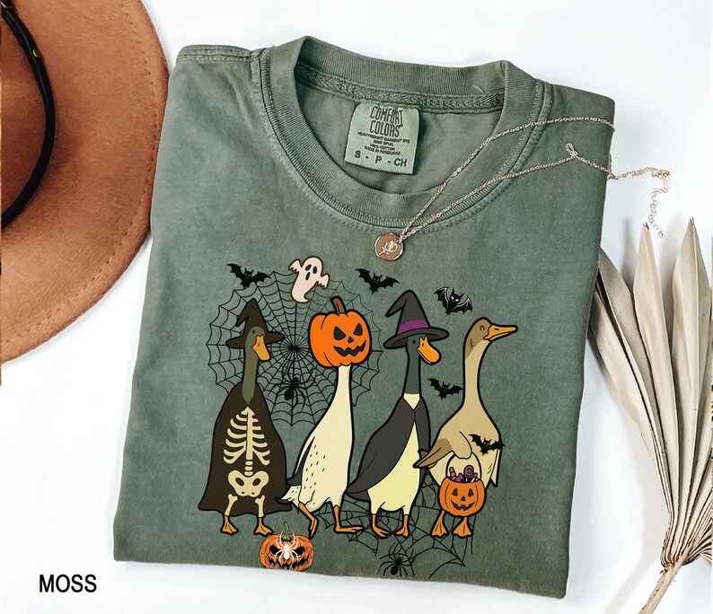 Puede incluir: Camiseta Comfort Colors verde musgo con un dise&ntilde;o de Halloween. El dise&ntilde;o incluye cuatro patos disfrazados, una telara&ntilde;a, murci&eacute;lagos y un fantasma. Un collar plateado est&aacute; sobre la camiseta.