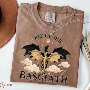 May include: Espresso-coloured t-shirt with a dragon graphic. The design features a dragon with black and gold wings, the text "FLY OR DIE" and "BASGIATH WAR COLLEGE". A silver necklace rests on the shirt.