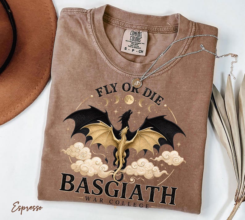 May include: Espresso-coloured t-shirt with a dragon graphic. The design features a dragon with black and gold wings, the text "FLY OR DIE" and "BASGIATH WAR COLLEGE". A silver necklace rests on the shirt.