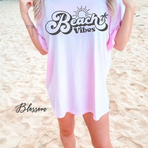 May include: A pink tie-dye t-shirt with the text "Beach Vibes" in a retro font. The text is in black and features a sun and palm tree graphic.