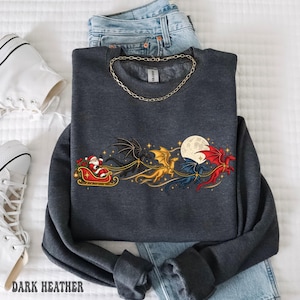 May include: A dark gray crewneck sweatshirt featuring a graphic print of Santa Claus in a sleigh pulled by three dragons in black, gold, and red, flying past a full moon. The sweatshirt is layered over blue denim jeans and a gold chain necklace.