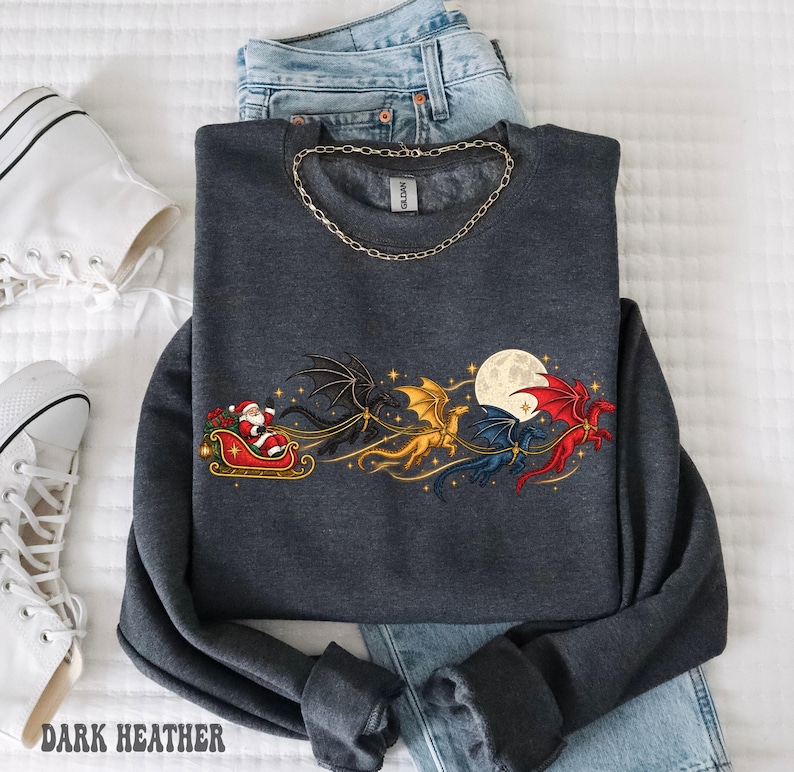 May include: A dark gray crewneck sweatshirt featuring a graphic print of Santa Claus in a sleigh pulled by three dragons in black, gold, and red, flying past a full moon. The sweatshirt is layered over blue denim jeans and a gold chain necklace.
