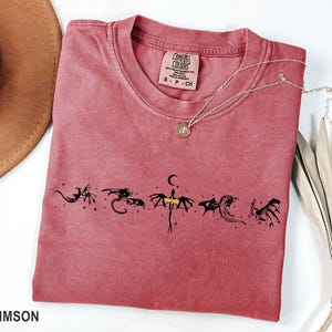 May include: Crimson-colored t-shirt with a crew neck and short sleeves. The shirt features a black dragon graphic design across the front. A silver necklace with a pendant is draped on the shirt. The tag reads "Comfort Colors".