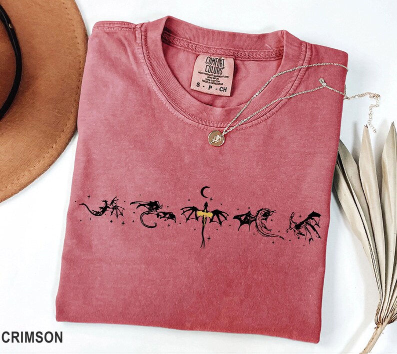 May include: Crimson-colored t-shirt with a crew neck and short sleeves. The shirt features a black dragon graphic design across the front. A silver necklace with a pendant is draped on the shirt. The tag reads "Comfort Colors".