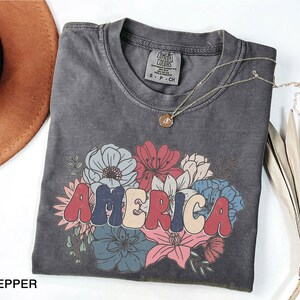 May include: A gray t-shirt with a vintage-style graphic of the word "AMERICA" in red, white, and blue letters surrounded by colorful flowers.