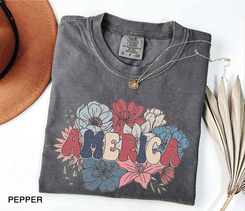 May include: A gray t-shirt with a vintage-style graphic of the word "AMERICA" in red, white, and blue letters surrounded by colorful flowers.