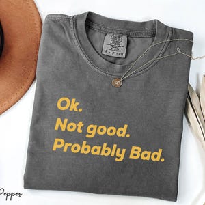 May include: A gray Comfort Colors t-shirt with the text "Ok. Not good. Probably Bad." in yellow. The shirt is folded and has a small tag at the neckline. A gold necklace with a pendant is draped across the shirt.