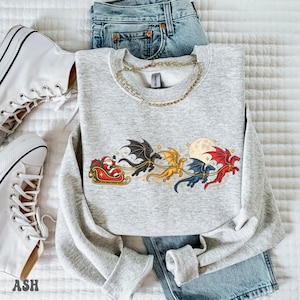 May include: A light gray crewneck sweatshirt featuring a graphic of Santa Claus in a red sleigh pulled by four flying dragons in black, gold, blue, and red colors, set against a full moon and small stars, paired with blue denim jeans.