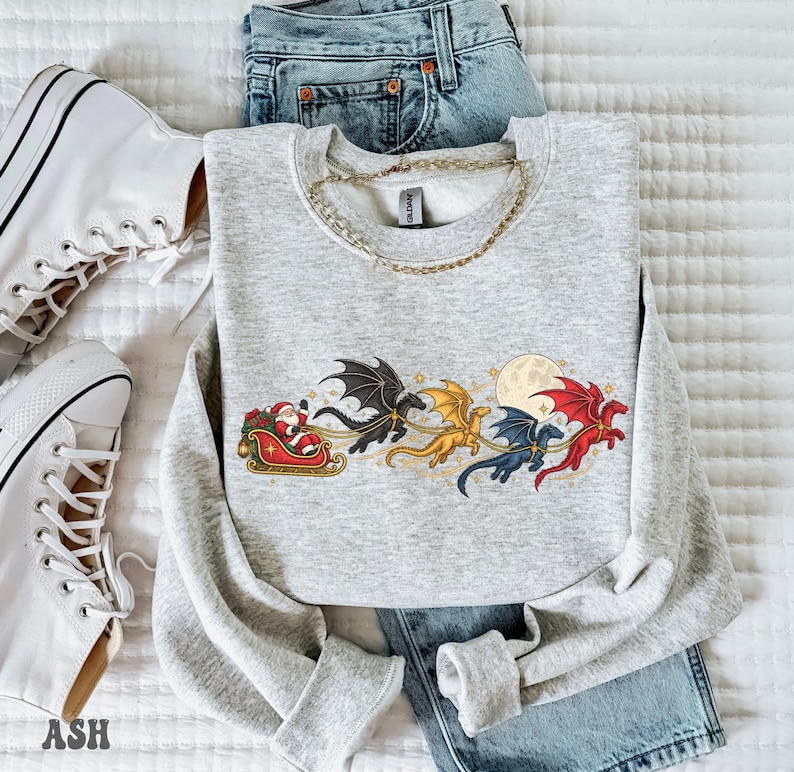 May include: A light gray crewneck sweatshirt featuring a graphic of Santa Claus in a red sleigh pulled by four flying dragons in black, gold, blue, and red colors, set against a full moon and small stars, paired with blue denim jeans.