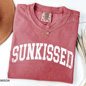May include: Crimson-colored t-shirt with the word "SUNKISSED" in white, block letters. A gold-colored necklace with a pendant rests on the shirt. The shirt is folded and placed on a white surface.