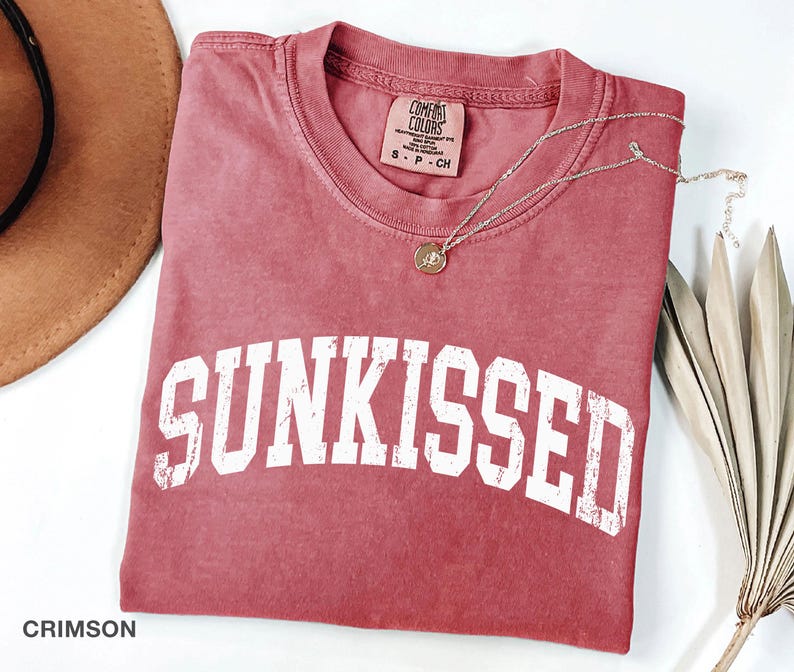 May include: Crimson-colored t-shirt with the word "SUNKISSED" in white, block letters. A gold-colored necklace with a pendant rests on the shirt. The shirt is folded and placed on a white surface.