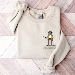 May include: A beige sweatshirt featuring a cartoon frog wearing a cowboy hat and holding a gun. The text "HOLD ON PARTNER IM OVERSTIMULAT" is printed below the frog.