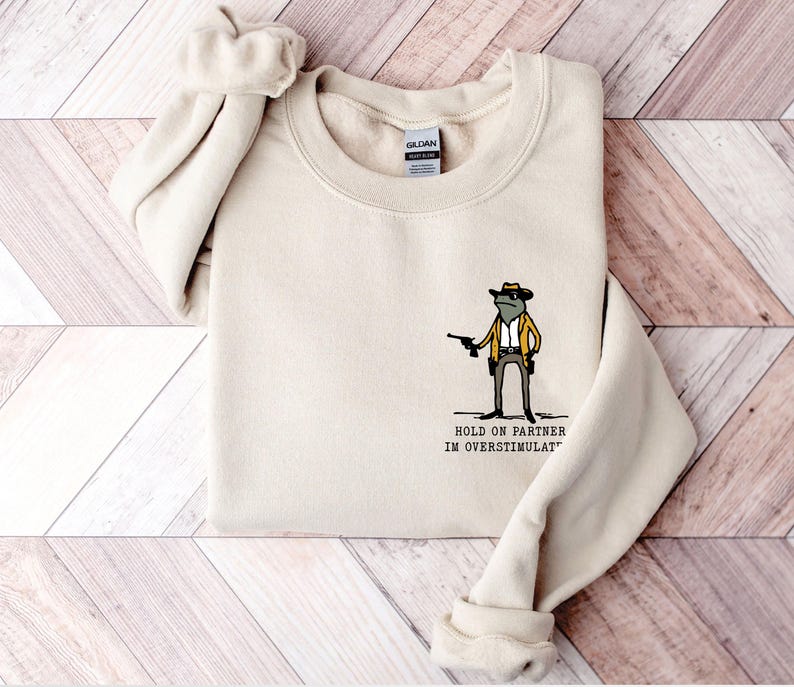 May include: A beige sweatshirt featuring a cartoon frog wearing a cowboy hat and holding a gun. The text "HOLD ON PARTNER IM OVERSTIMULAT" is printed below the frog.