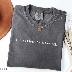 May include: A dark gray t-shirt with the text "I'd Rather Be Reading." in white lettering. The shirt is made of a soft, comfortable material and has a vintage look.