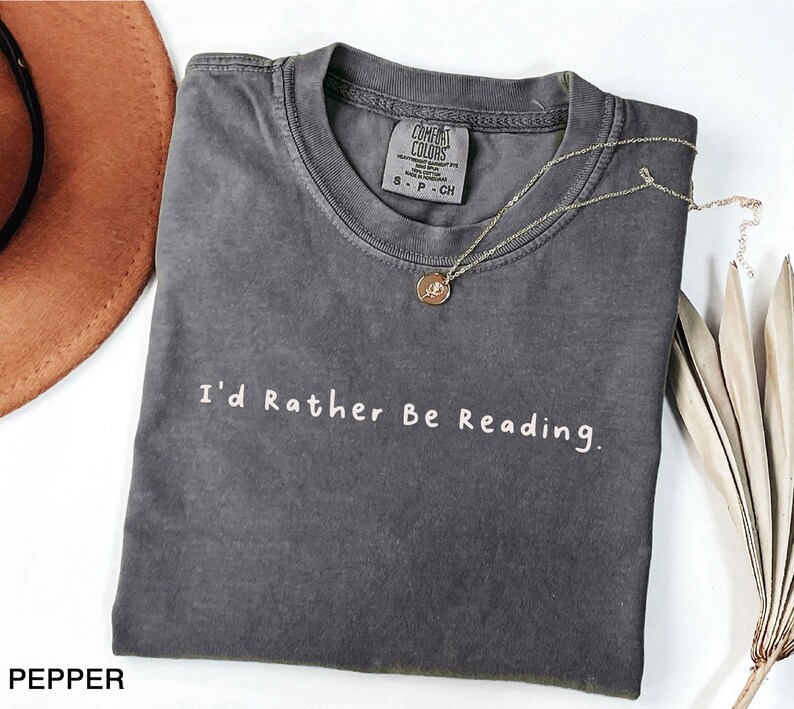 May include: A dark gray t-shirt with the text "I'd Rather Be Reading." in white lettering. The shirt is made of a soft, comfortable material and has a vintage look.