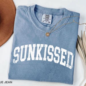 May include: A blue jean colored t-shirt with the word "SUNKISSED" in white block letters. A gold necklace with a pendant rests on the shirt. The shirt is folded, and a brown hat is visible in the background.