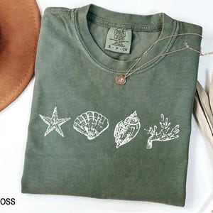 Op de afbeelding: Een mosgroen T-shirt met een wit ontwerp van een zeester, schelpen en koraal. Het shirt heeft de tekst "COMFORT COLORS" en "S - P - CH" op het label.
