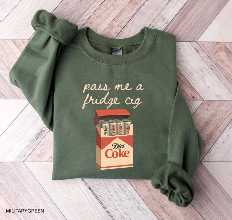 May include: Military green sweatshirt with the phrase "pass me a fridge cig" in a handwritten font. The graphic features a Diet Coke cigarette box with Diet Coke cans inside.