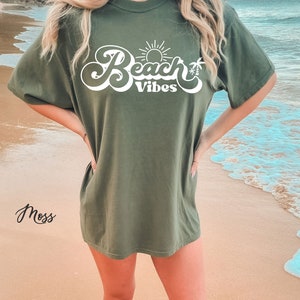 May include: A green oversized t-shirt with the text "Beach Vibes" in white cursive lettering. The text includes a sun and palm tree graphic.