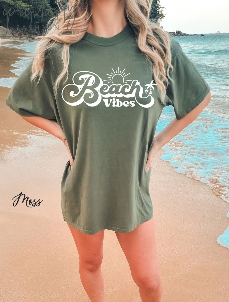 May include: A green oversized t-shirt with the text "Beach Vibes" in white cursive lettering. The text includes a sun and palm tree graphic.