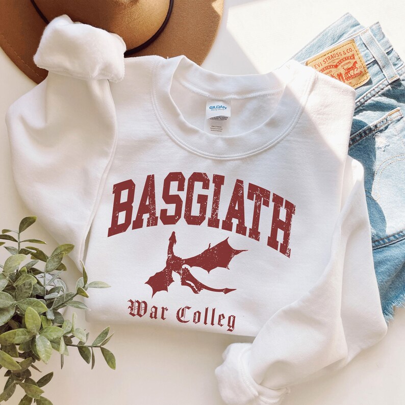 May include: White crewneck sweatshirt with the word "BASGIATH" in distressed red lettering, a red dragon graphic, and the words "War Colleg". The sweatshirt is folded with a pair of denim shorts and a brown hat in the background.