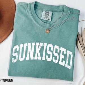 May include: A light green t-shirt with the word "SUNKISSED" in large white block letters. A silver necklace with a small pendant rests on the shirt. The shirt is folded and displayed with a brown hat and dried plant.