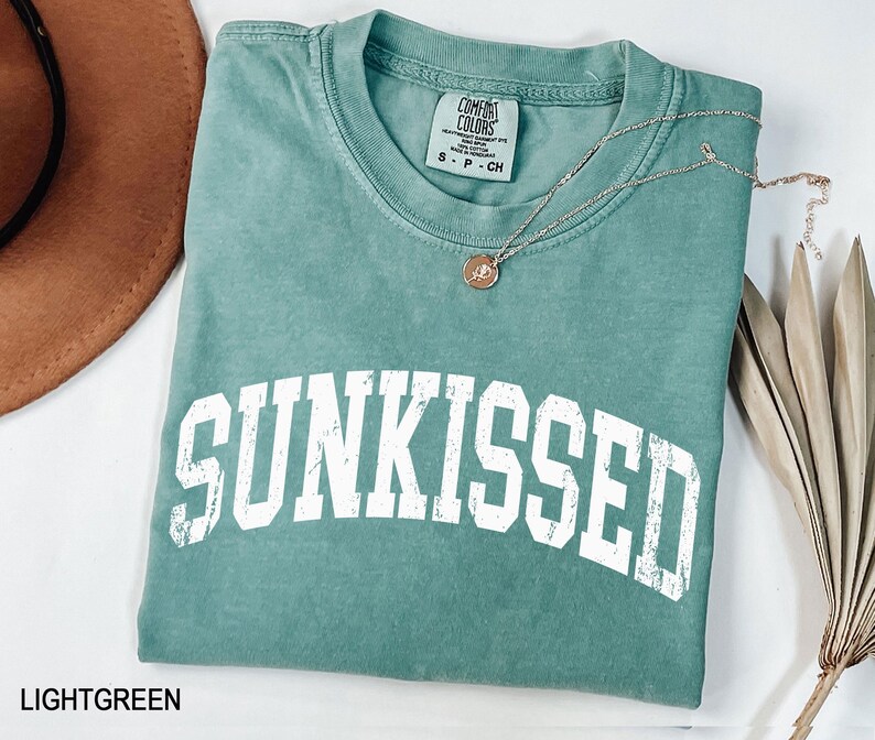 May include: A light green t-shirt with the word "SUNKISSED" in large white block letters. A silver necklace with a small pendant rests on the shirt. The shirt is folded and displayed with a brown hat and dried plant.