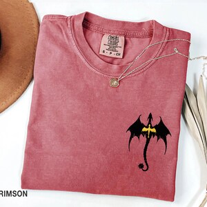 May include: Crimson red t-shirt with a black dragon silhouette with yellow accents. The shirt is made by Comfort Colors and has a tag that says "S - P - CH".