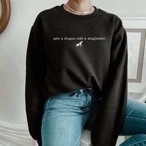May include: Black sweatshirt with the text "save a dragon ride a wingleader." and a small graphic of a dragon. The model is wearing blue jeans.