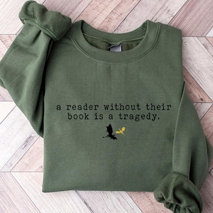 May include: A dark green sweatshirt with the text "a reader without their book is a tragedy." printed on the front. The text is in black and there is a black silhouette of a dragon and a gold silhouette of a dragon above the text.