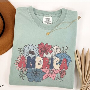 May include: A light blue t-shirt with a graphic of the word "AMERICA" in colorful, retro-style letters surrounded by a floral design. The flowers are red, blue, pink, and white.