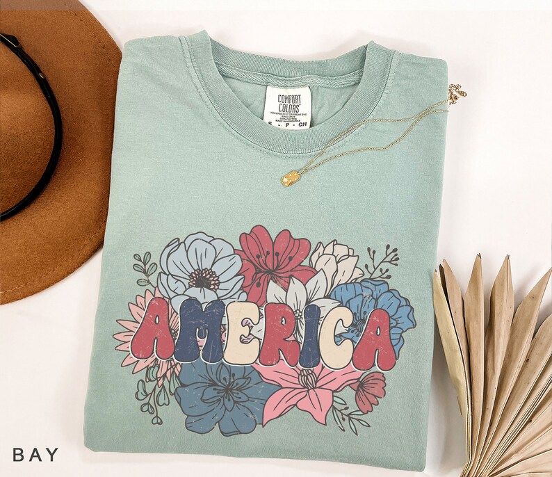 May include: A light blue t-shirt with a graphic of the word "AMERICA" in colorful, retro-style letters surrounded by a floral design. The flowers are red, blue, pink, and white.