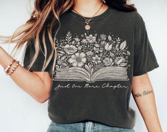 May include: A dark grey t-shirt featuring a graphic of an open book with flowers growing out of it. The text "Just One More Chapter" is printed below the book.