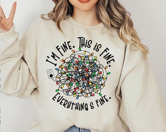 May include: Beige sweatshirt with the text "I'm Fine. This is Fine. Everything's Fine." and a graphic of tangled Christmas lights. The design includes a power outlet. A casual, comfortable top.