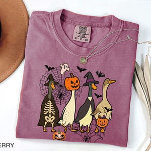 Puede incluir: Camiseta color bayas con un dise&ntilde;o de Halloween de cuatro patos disfrazados. Un pato est&aacute; vestido de esqueleto, otro de bruja, un tercero con cabeza de calabaza y el &uacute;ltimo sosteniendo una calabaza. La camiseta tiene cuello redondo y una etiqueta Comfort Colors.