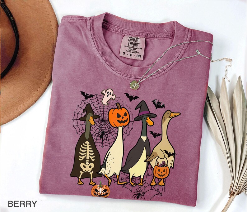 Puede incluir: Camiseta color bayas con un dise&ntilde;o de Halloween de cuatro patos disfrazados. Un pato est&aacute; vestido de esqueleto, otro de bruja, un tercero con cabeza de calabaza y el &uacute;ltimo sosteniendo una calabaza. La camiseta tiene cuello redondo y una etiqueta Comfort Colors.