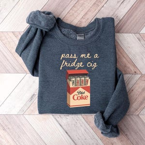 May include: A navy blue sweatshirt with the text "pass me a fridge cig" in a cursive font. Below the text is a graphic of a Diet Coke box with Diet Coke cans inside.