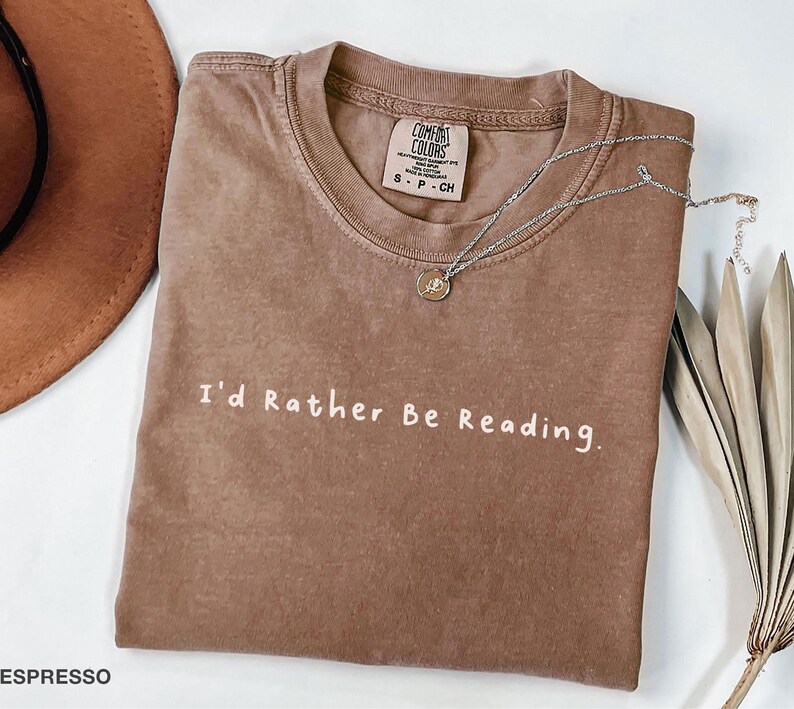 May include: A brown t-shirt with the text "I'd Rather Be Reading." printed in white. The shirt is made of a soft, comfortable material and has a relaxed fit.