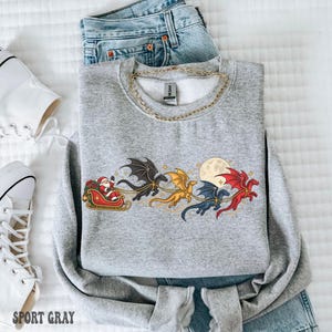 May include: A heather gray crewneck sweatshirt featuring a graphic print of Santa Claus in a red sleigh pulled by four colorful dragons in black, gold, blue, and red, flying past a full moon. The garment has a gold chain necklace resting on top.