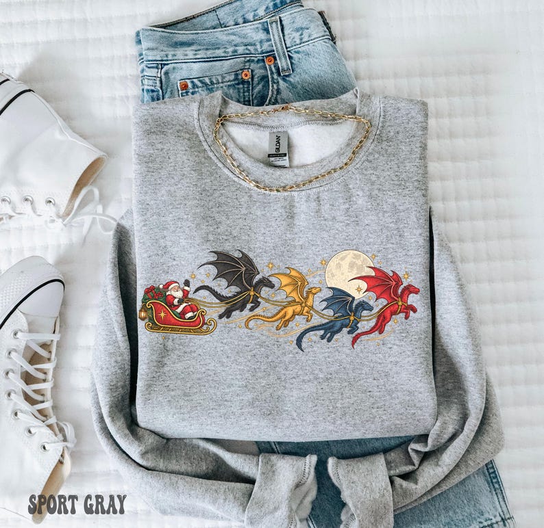 May include: A heather gray crewneck sweatshirt featuring a graphic print of Santa Claus in a red sleigh pulled by four colorful dragons in black, gold, blue, and red, flying past a full moon. The garment has a gold chain necklace resting on top.