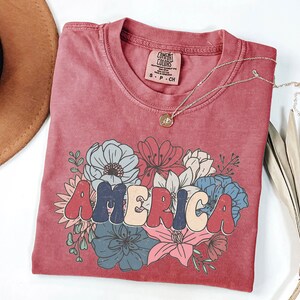 May include: A red t-shirt with a vintage-style graphic of the word "AMERICA" surrounded by colorful flowers. The shirt is made of a soft, comfortable material and has a distressed look.