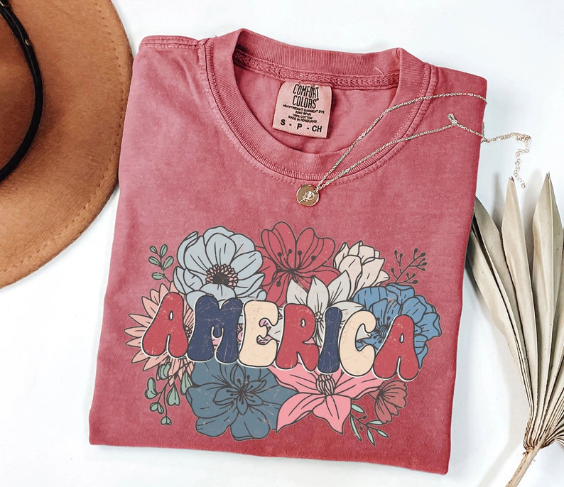 May include: A red t-shirt with a vintage-style graphic of the word "AMERICA" surrounded by colorful flowers. The shirt is made of a soft, comfortable material and has a distressed look.