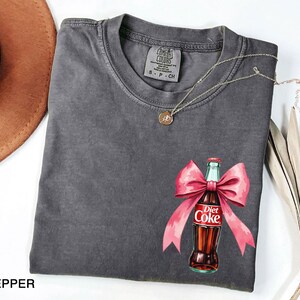 May include: A gray t-shirt with a pink bow and a Diet Coke bottle graphic. The text on the bottle reads "Diet Coke".