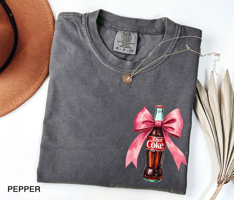 May include: A gray t-shirt with a pink bow and a Diet Coke bottle graphic. The text on the bottle reads "Diet Coke".